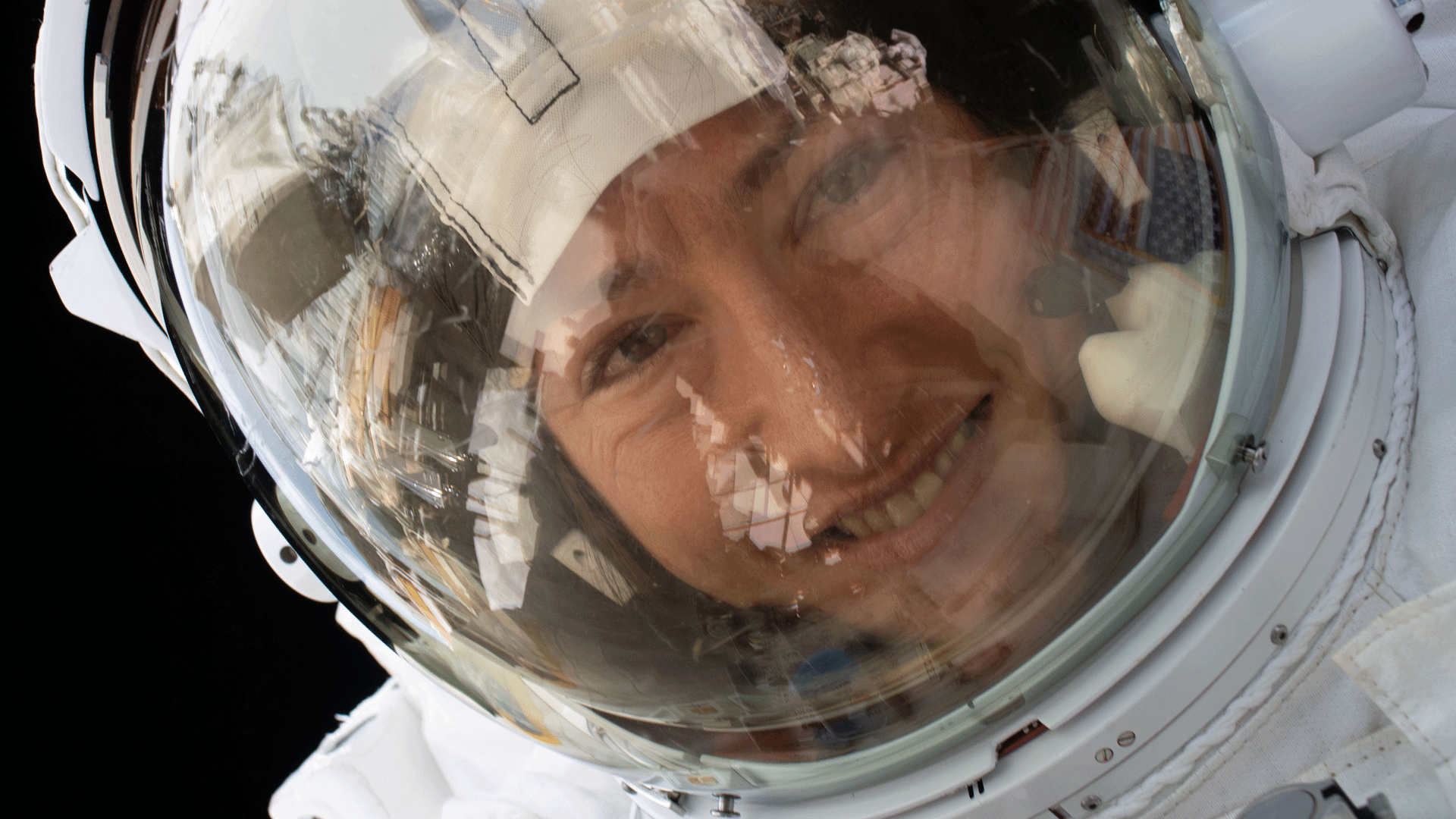 Christina Koch smiles from behind a sealed space suit visor. The reflection of the International Space Station and Earth can be seen in reflection on the glass