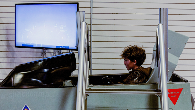 A boy with messy, curly hair sits in the cockpit of an airplane simulator. The screen showing his view displays a clear blue sky.