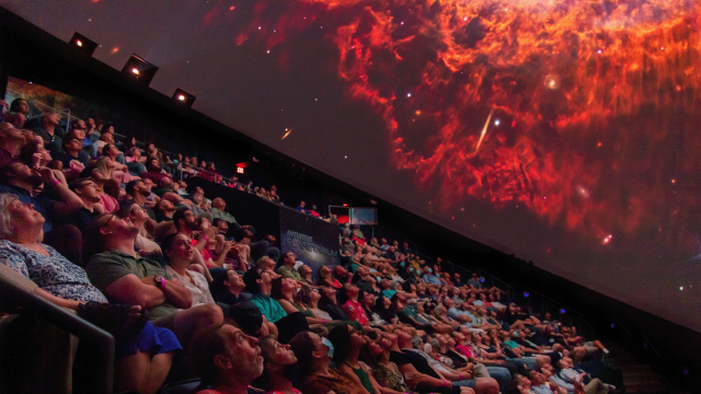 An audience sits in the dark INTUITIVE Planetarium looking up at an image of The Southern Ring Nebula