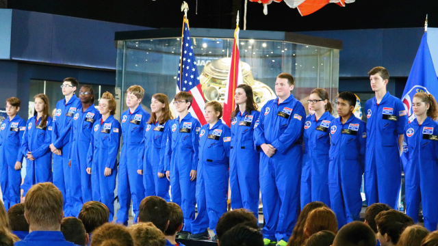 Space Camp graduates in blue uniforms stand in front of the Apollo 16 capsule