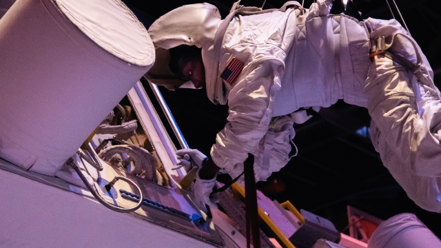 a trainee in a white extravehicular spacesuit and helmet is suspended midair on a harness to simulate satellite repair 