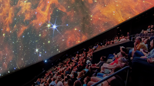 A crowd looks up at an image of a galaxy displayed on a large dome screen overhead