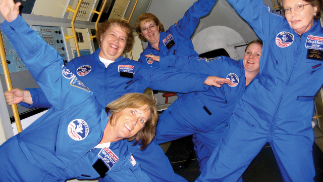 A group of educators in blue flight suits pose with their arms out as if in zero gravity