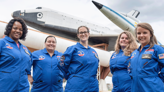 A group of trainees in blue flight suits pose in front of the Space Shuttle Pathfinder and the T-38