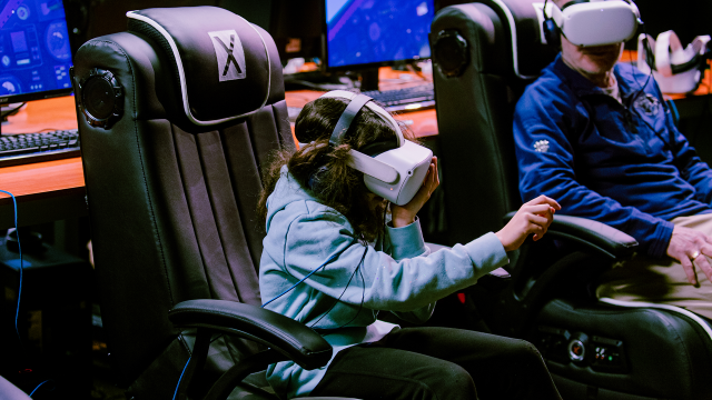 A young guests wearing a headset covering her eyes and ears, reaches out to touch buttons she can see inside a virtual Apollo 11 capsule