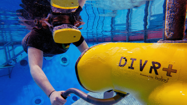 A swimmer at the surface of the water wears a yellow virtual reality helmet while gripping a yellow stationary thruster emits haptic feedback synced to the visuals