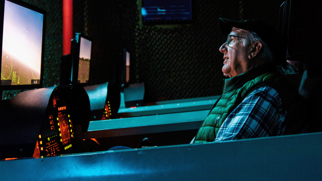 An older man sits in the Aviation Challenge flight simulator cockpit. The sky and distant horizon can be seen on the screen.