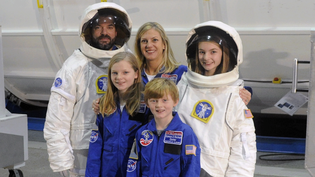 A family stands on the mission control floor some in helmets and white extravehicular spacesuits and others in blue flight suits.