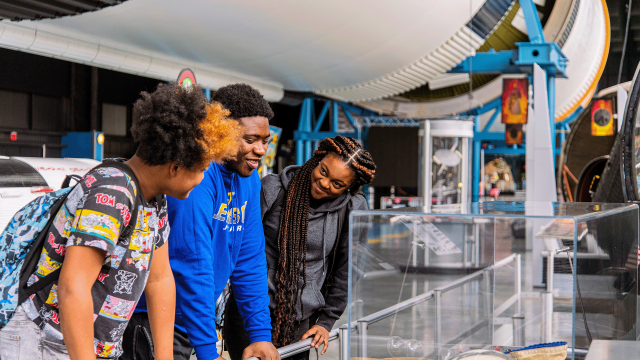 Three guests examine a display. The Saturn V launch vehicle is suspended overhead.