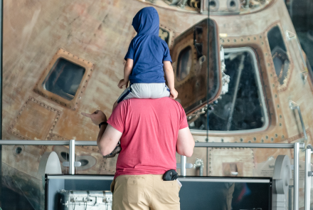 a child sits on their father's shoulders while they explore the Apollo 16 exhibit 