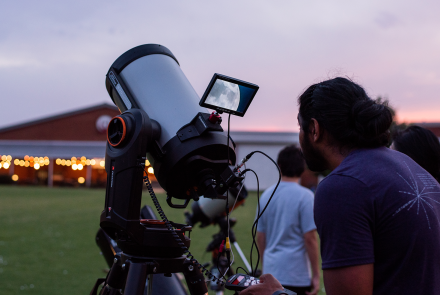 A telescope points to the sky, displaying an image of the moon on the screen to an interested viewer